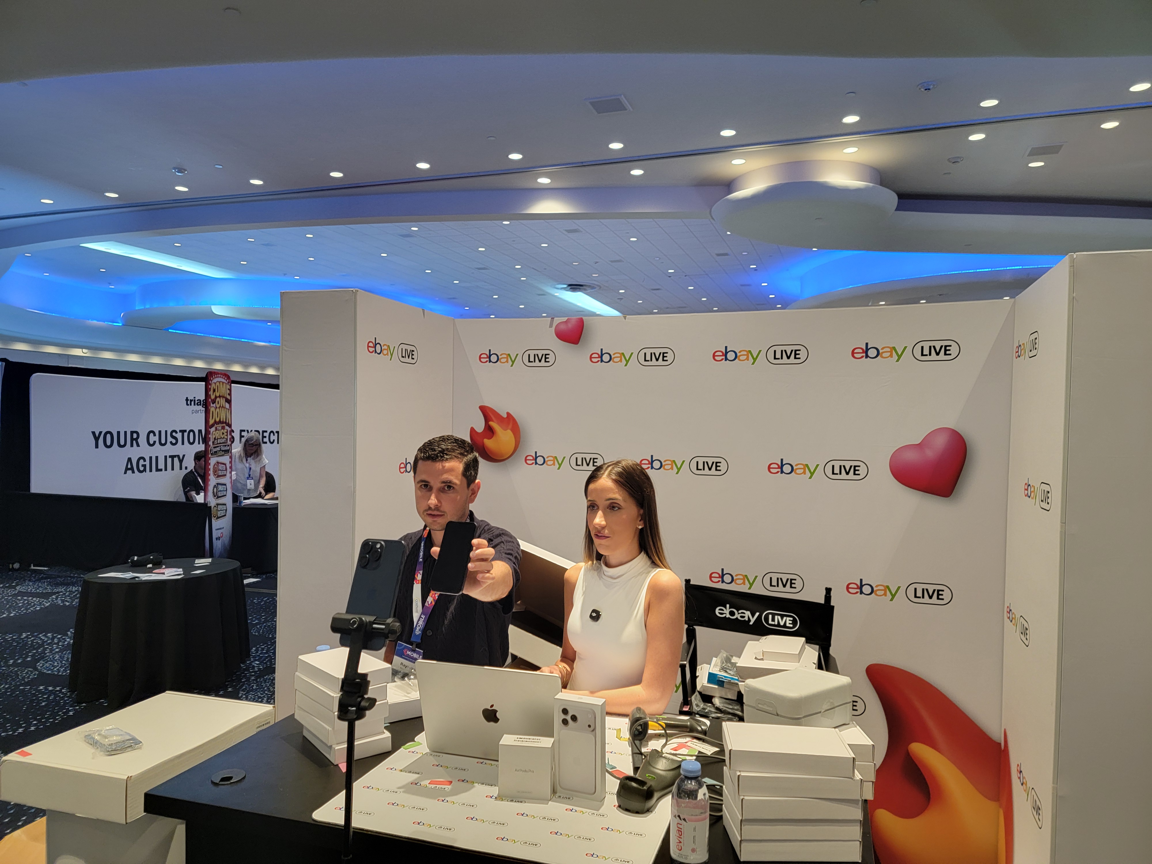 Two hosts live streaming at the eBay Live booth at Mobile Disrupt 2026, holding up smartphones on camera with stacks of boxed devices on the table